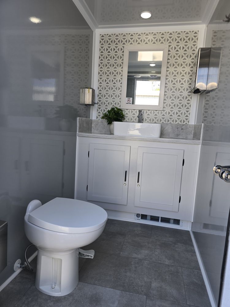 Interior of a portable restroom with a white toilet, sink, and patterned wall. Gray floor and cabinetry.