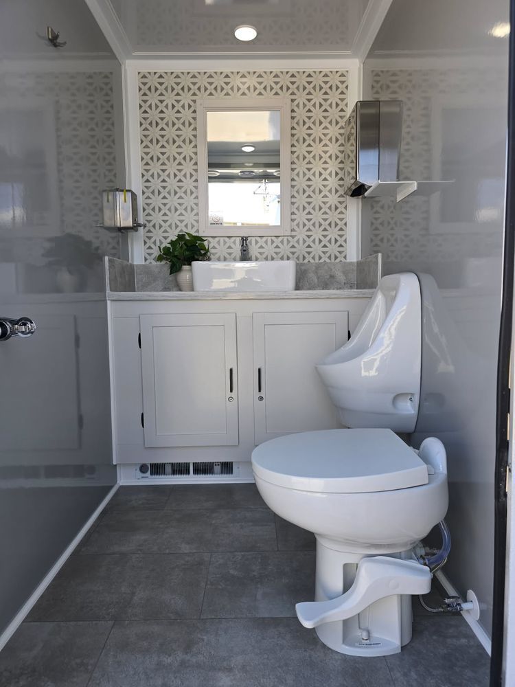 Interior view of a luxury portable restroom with a toilet, urinal, sink, and decorative tile.