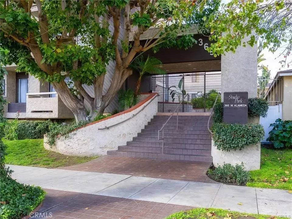 Apartment entrance with steps, white ramp wall, trees, and a black sign by the walkway