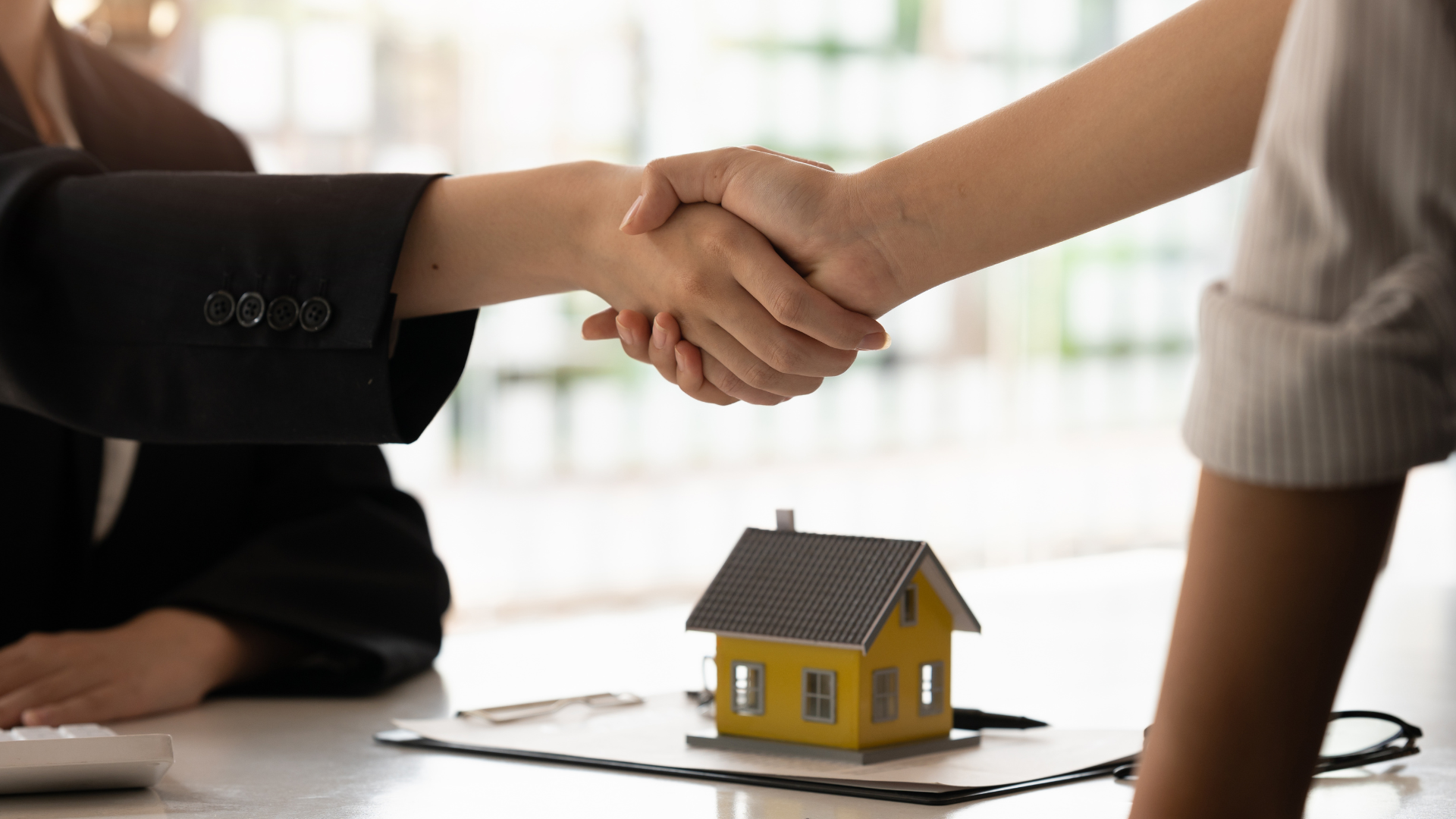 Two people shaking hands beside a small model house on a desk