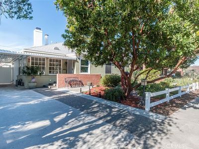 Front patio of a house with a large tree, white fence, and sunny driveway