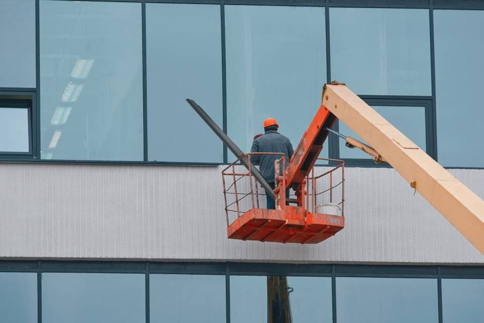 A worker in an orange hard hat standing in a red aerial lift platform, working on the exterior of a glass-paneled building.
