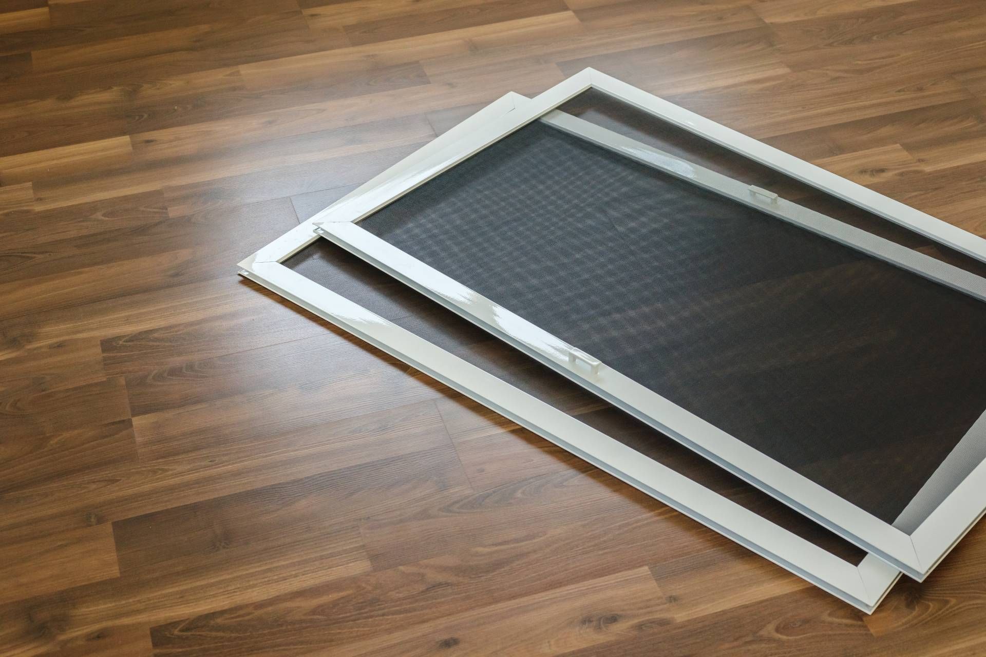Two white-framed window screens with dark mesh stacked on a brown wood-grain floor.