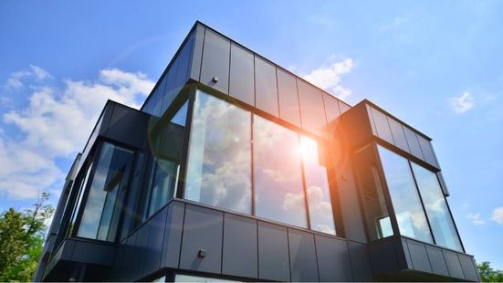 A modern, multi-story building with dark metal paneling and large floor-to-ceiling windows under a bright, sunny sky.