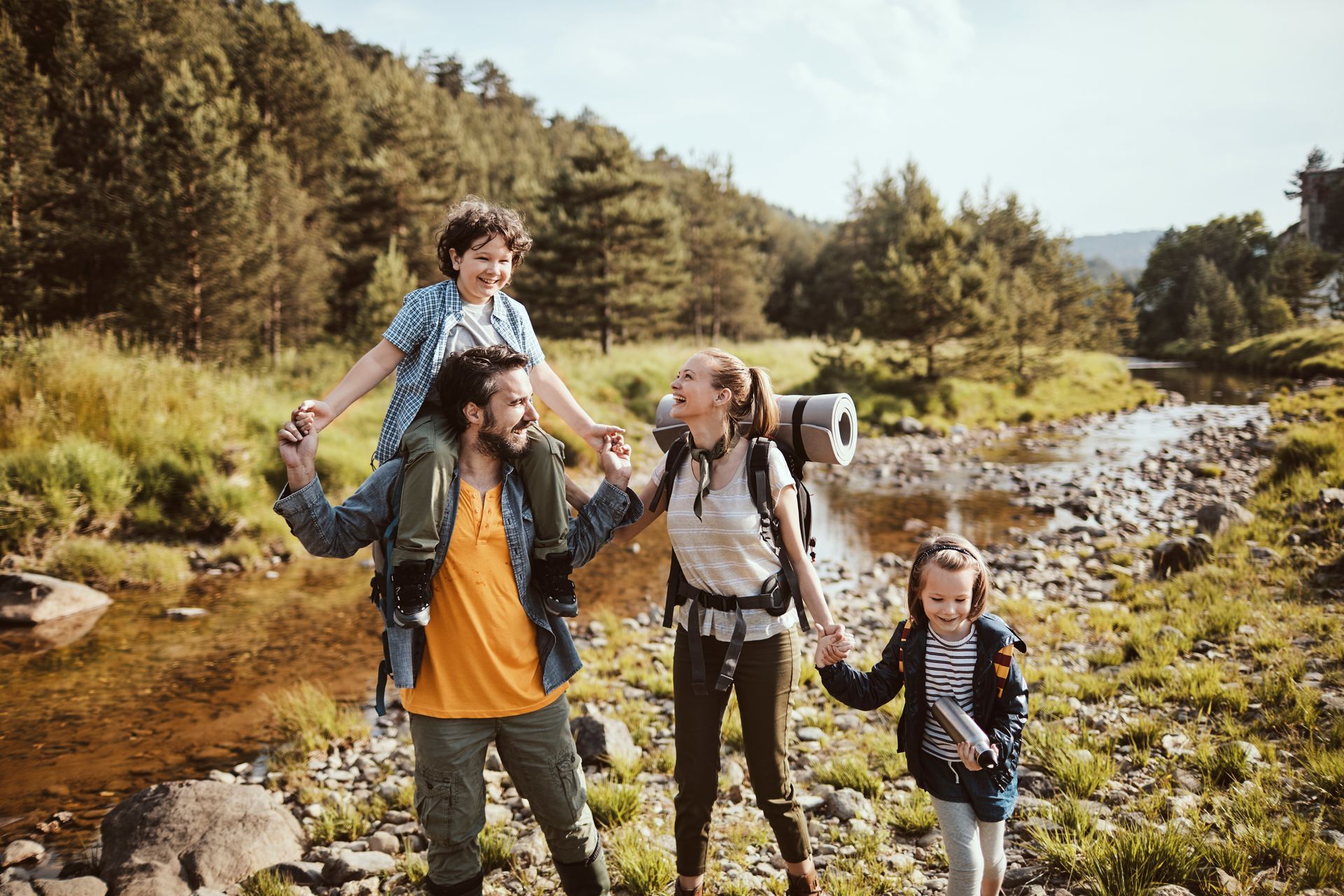 Family hiking along a rocky stream in a sunny forested valley, smiling and carrying a child on shoulders