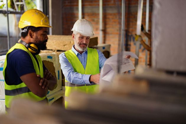 Two construction workers in hard hats and safety vests reviewing plans in a building site.