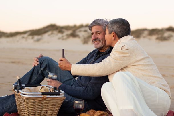 Two men sitting on a beach picnic blanket, smiling while sharing food and drinks at sunset