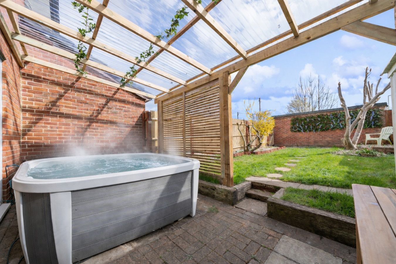 Covered backyard patio with a hot tub, wooden pergola, and grassy garden area.