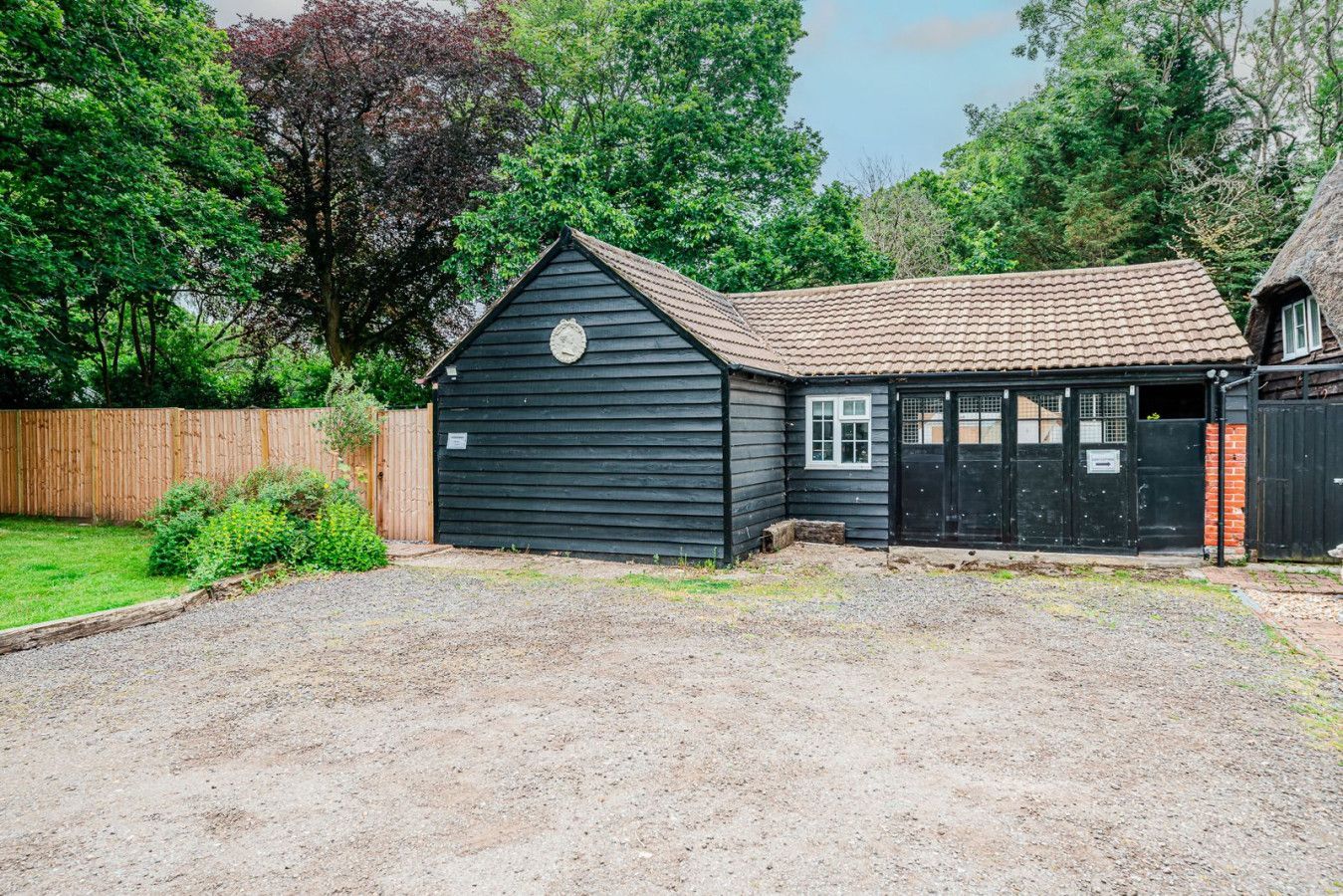 Small black wooden cottage with gravel driveway, surrounded by trees and a wooden fence.