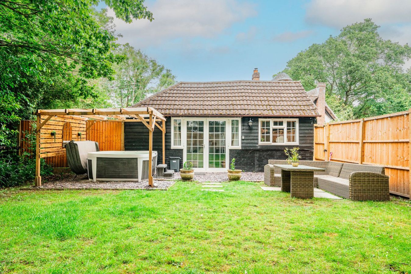 Backyard with lawn, patio seating, wooden pergola, fenced garden, and a small house in the background