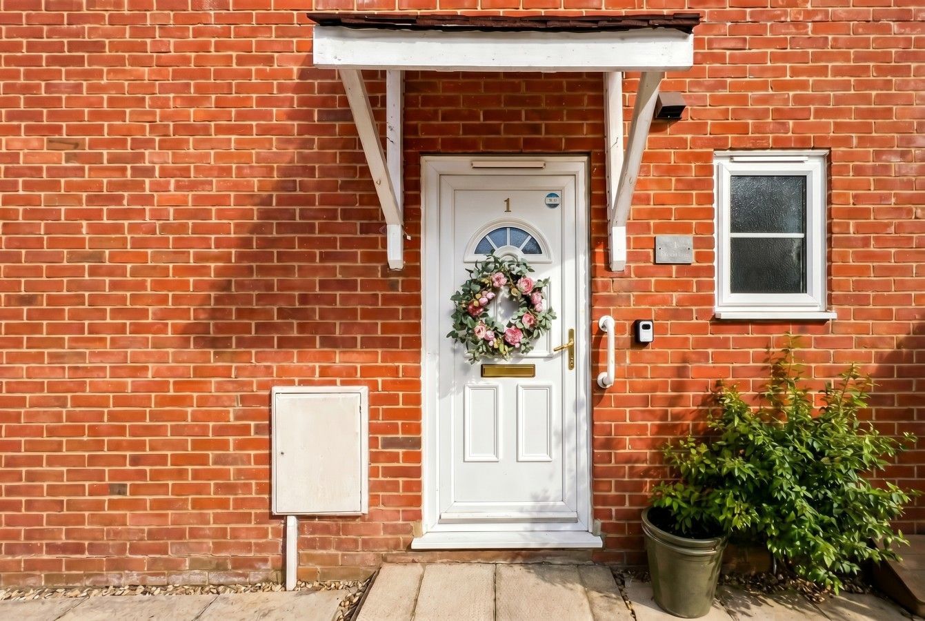 Front door of a red brick house with white trim, wreath, porch awning, window, and potted plant