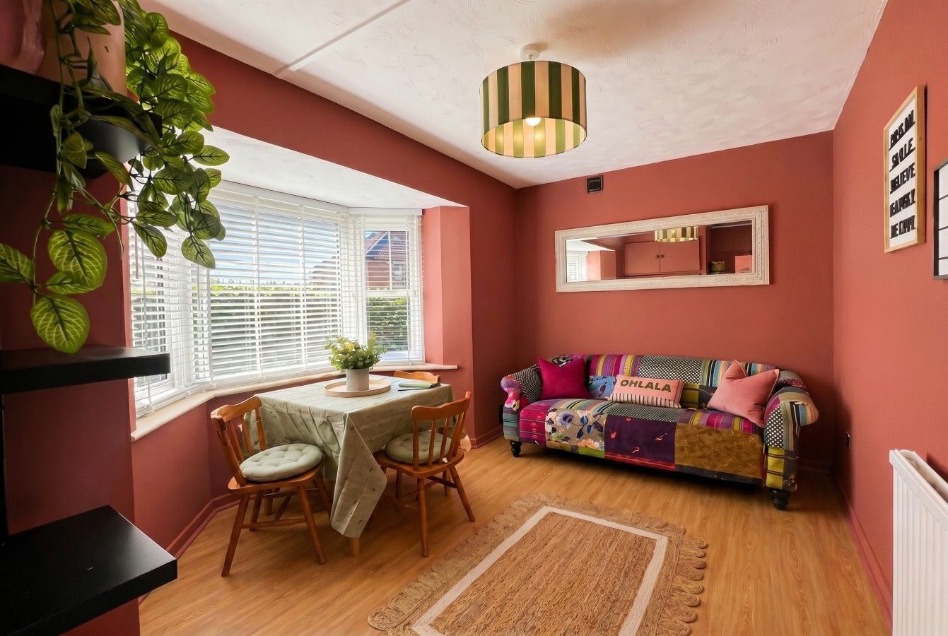 Cozy pink living room with a window bench, small dining table, colorful sofa, and hanging plants.