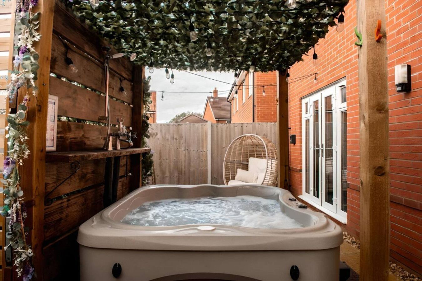 Covered hot tub in a narrow brick courtyard with string lights and a white chair at the far end