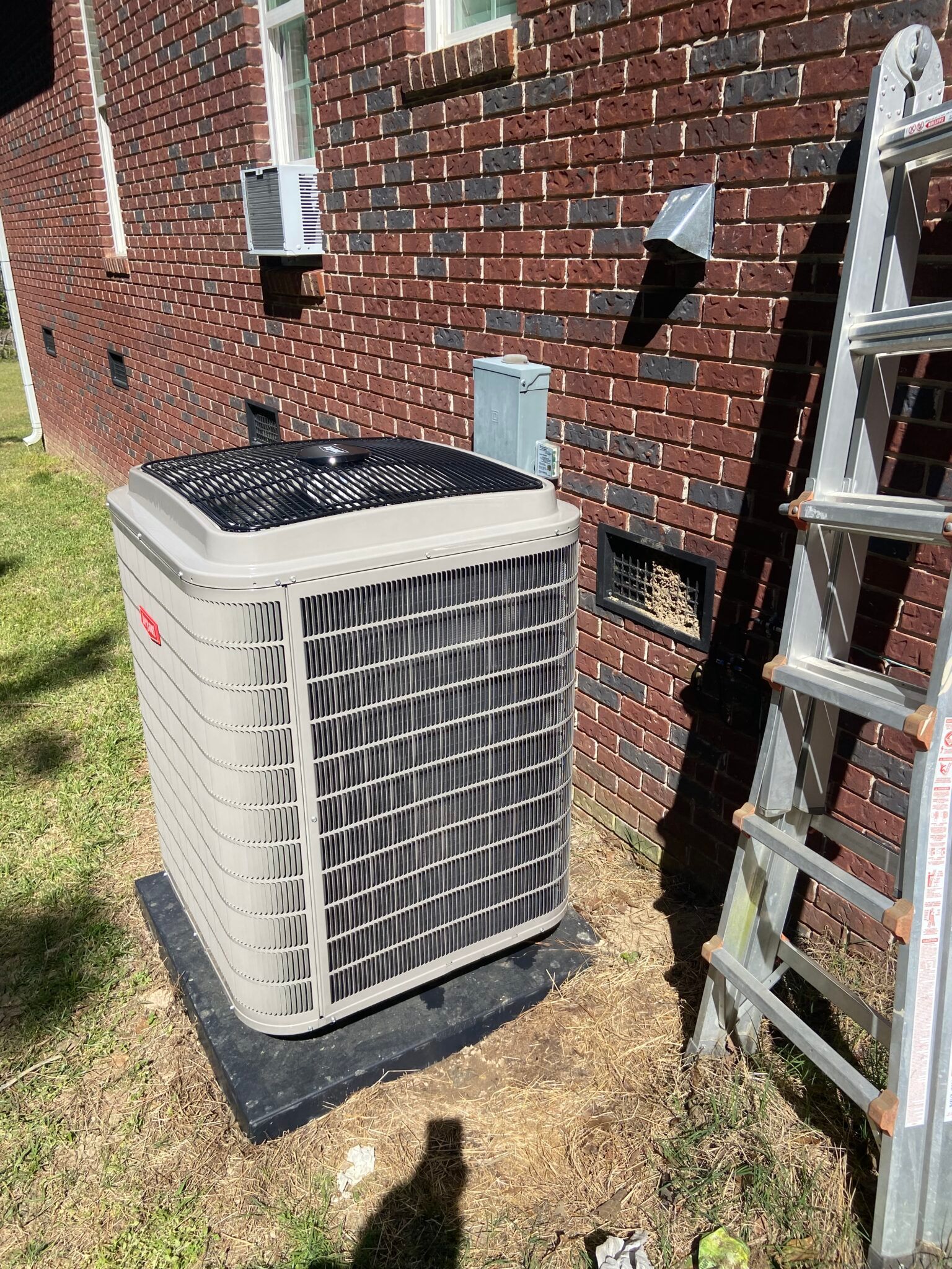 An air conditioner is sitting on the side of a brick building next to a ladder.