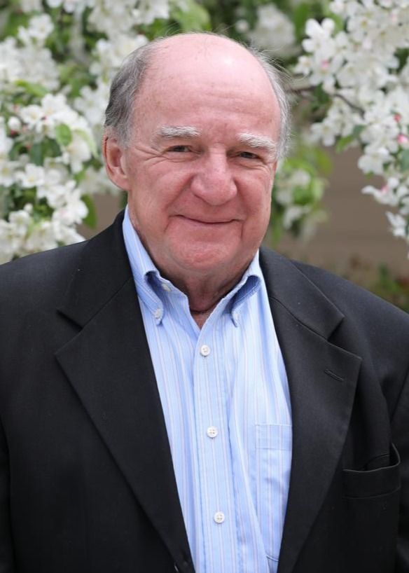 Man in a black blazer and blue shirt smiles in front of blooming white flowers.