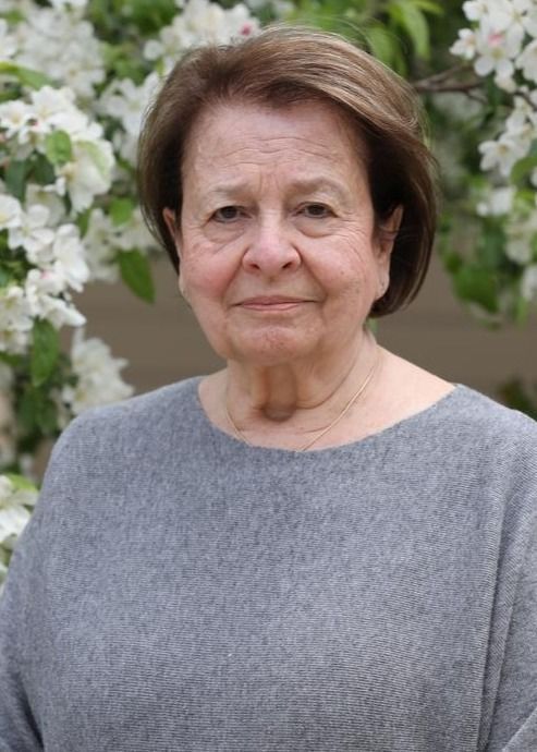 Woman with short brown hair in gray sweater, standing in front of blooming white flowers.