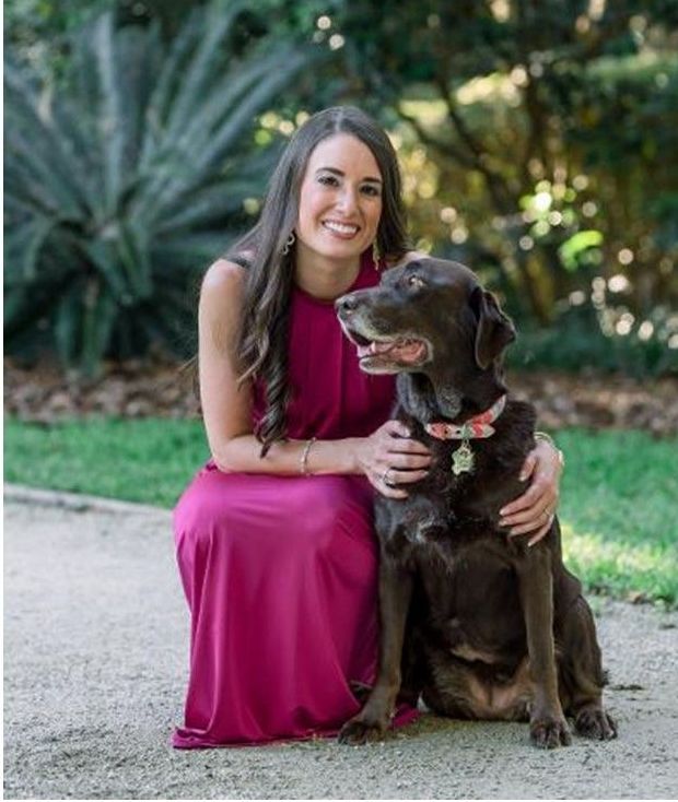 Woman in pink dress kneels with brown dog on a path in a garden, smiling.