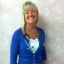 Smiling person with a stethoscope, wearing a blue cardigan over a white top, standing near a pink wall.