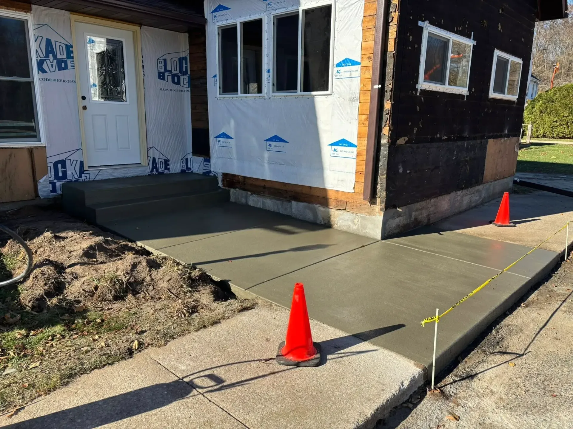 A new concrete patio with steps leading to a house under renovation, marked by orange safety cones and caution tape.