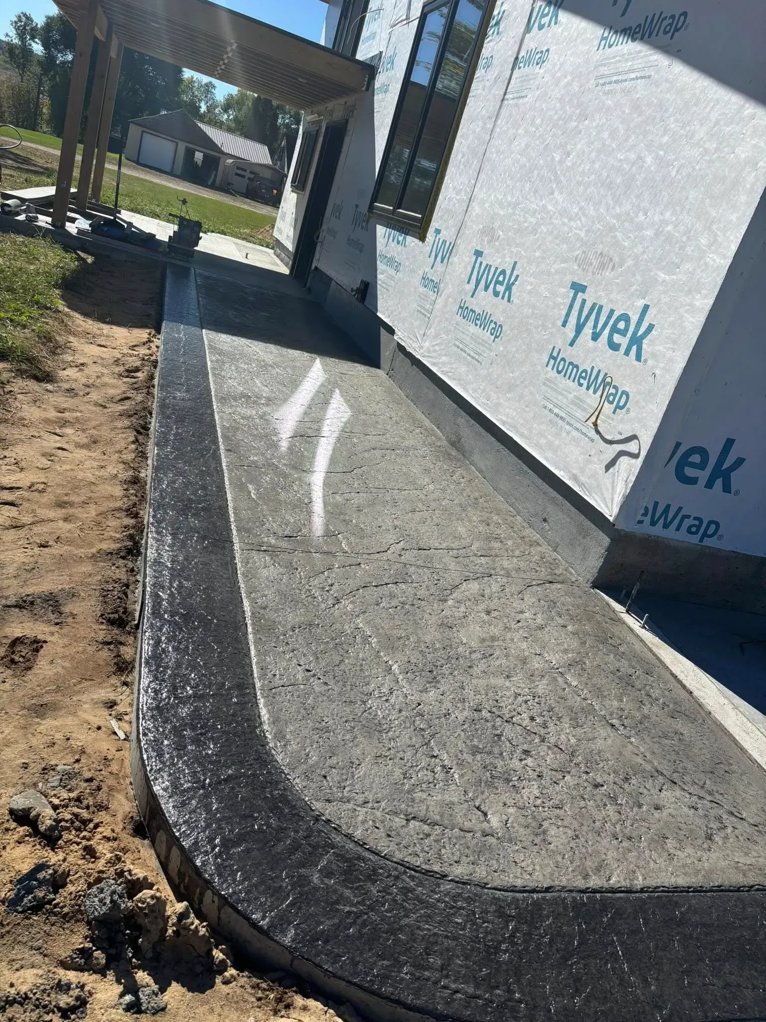 New concrete walkway with a dark border installed along the side of a building wrapped in Tyvek house wrap.