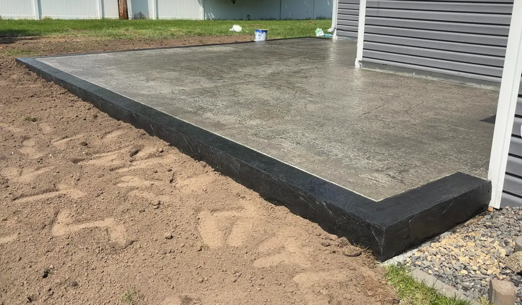 A newly poured concrete patio with a dark, textured border edge set against a house and an unfinished dirt yard.