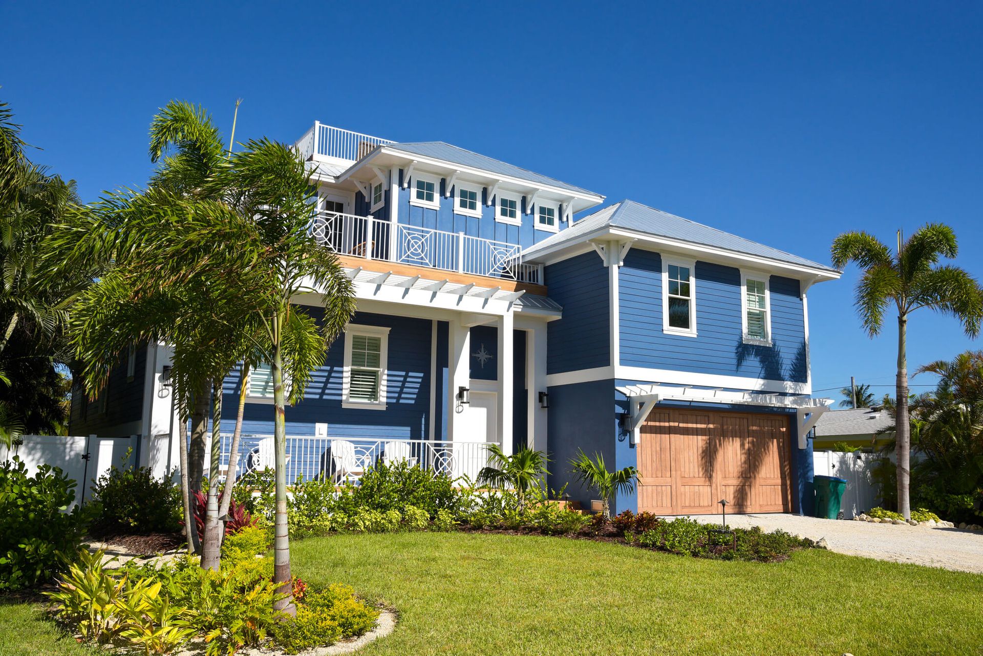 A blue two-story coastal house with a white trim, a covered porch, and a brown garage, set under a clear blue sky.