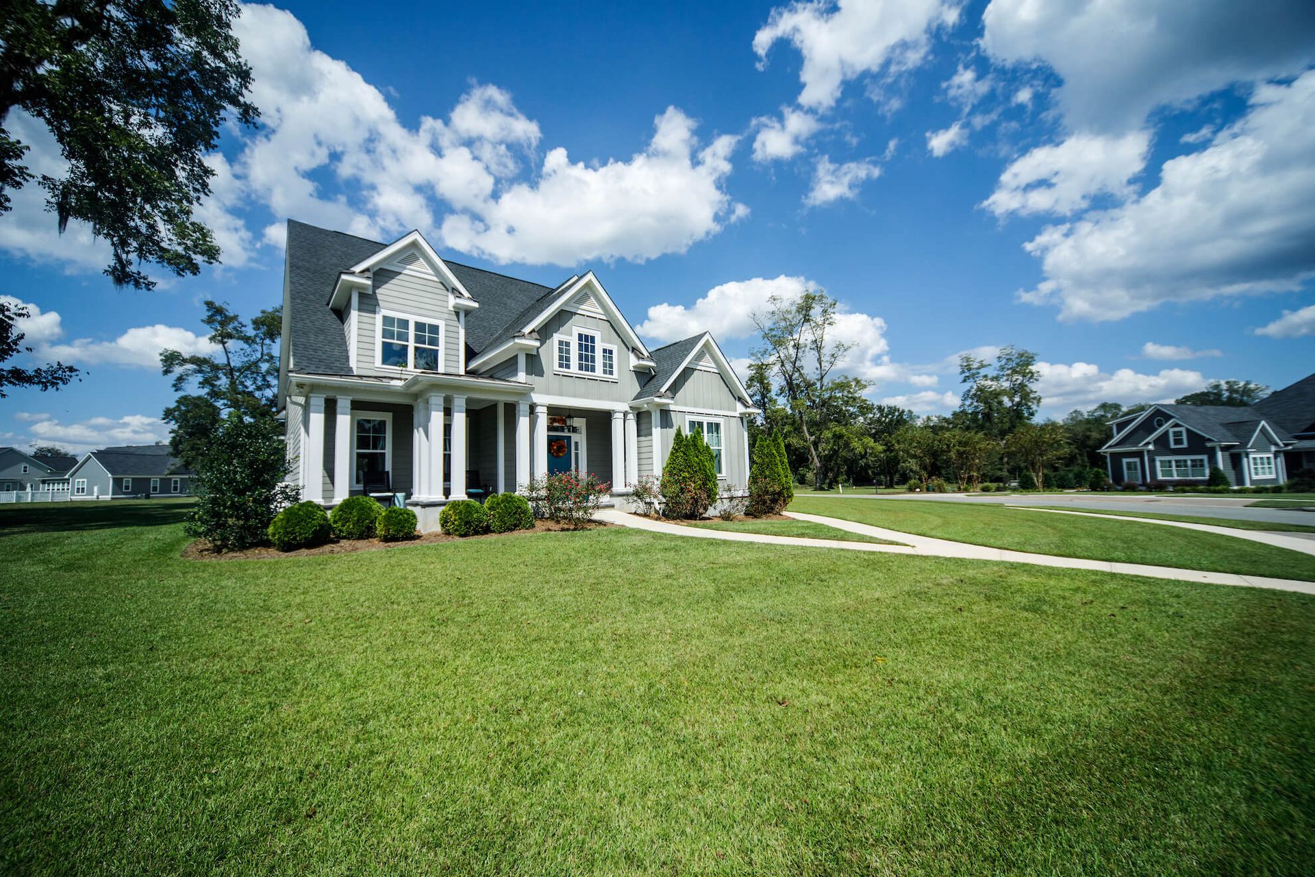 A two-story grey suburban house with a large green lawn under a bright blue, partly cloudy sky.