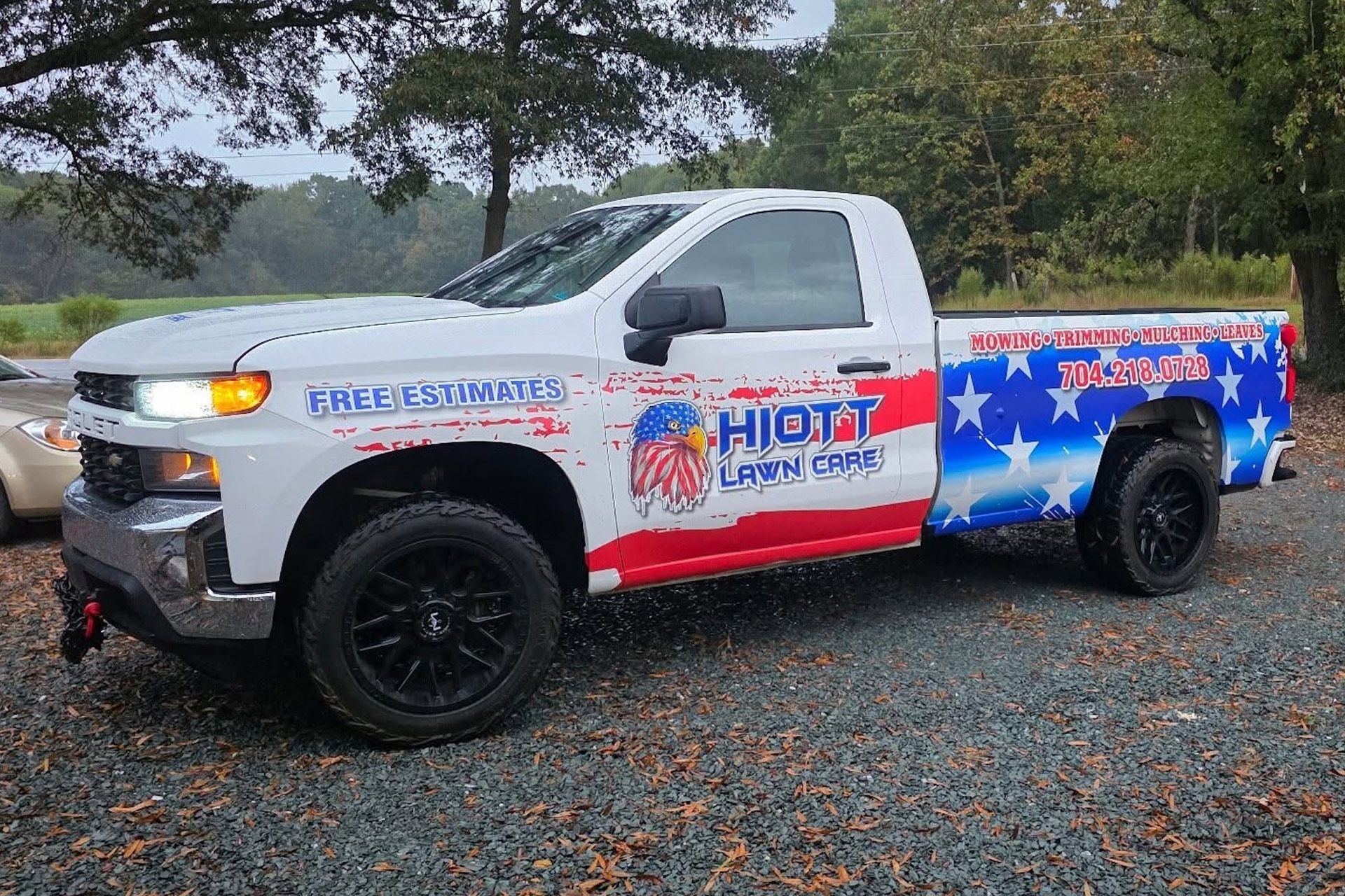 White pickup truck with red, white, and blue graphics for HIOTT Lawn Care parked on gravel.