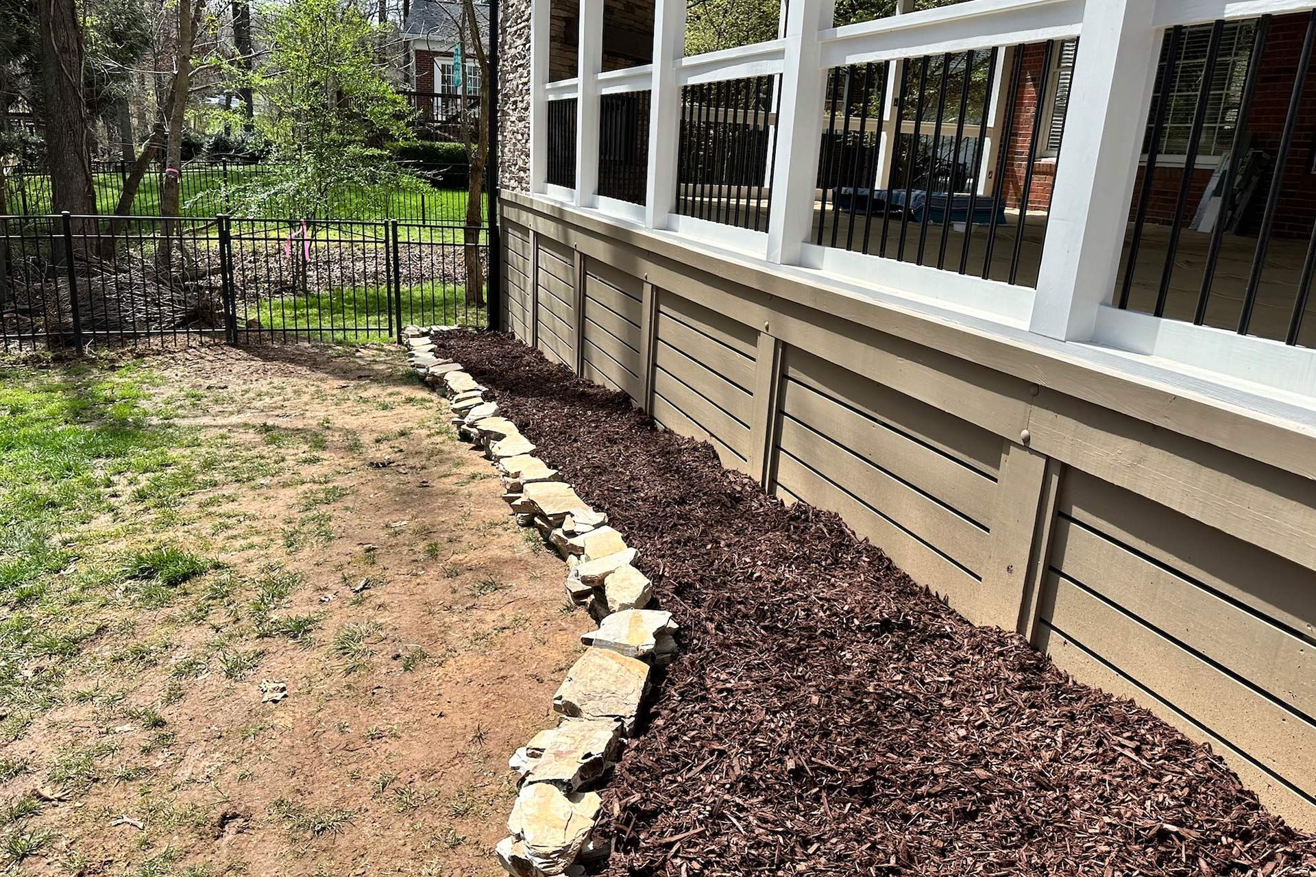 A garden bed with brown mulch along the side of a porch with a stone border.
