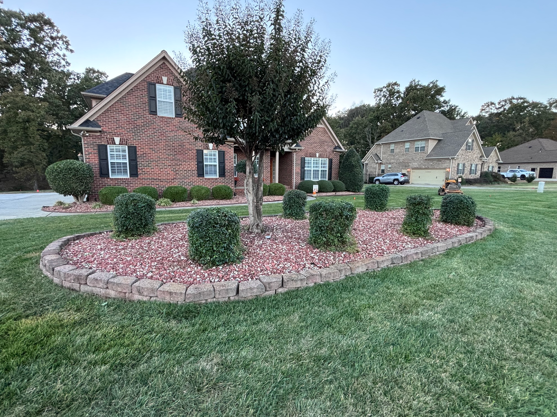 Brick house with landscaped front yard, a curved flower bed with red rocks, trimmed green bushes, and a tree.