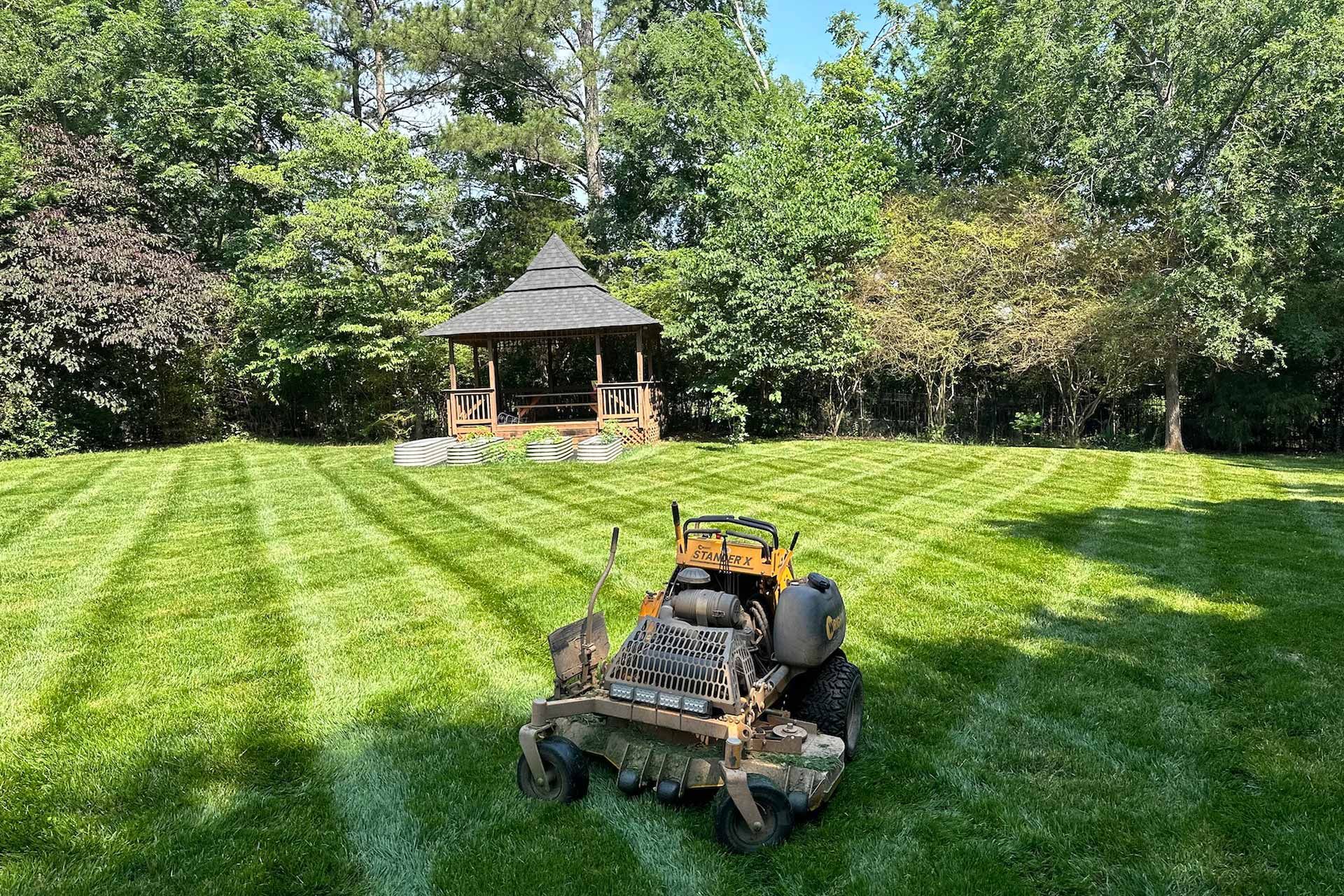 Lawn mower cutting grass in a backyard with a gazebo in the background on a sunny day.