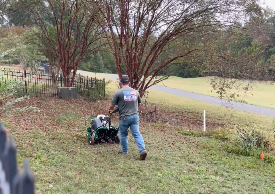 Man using a tiller on a hillside near trees and a golf course.