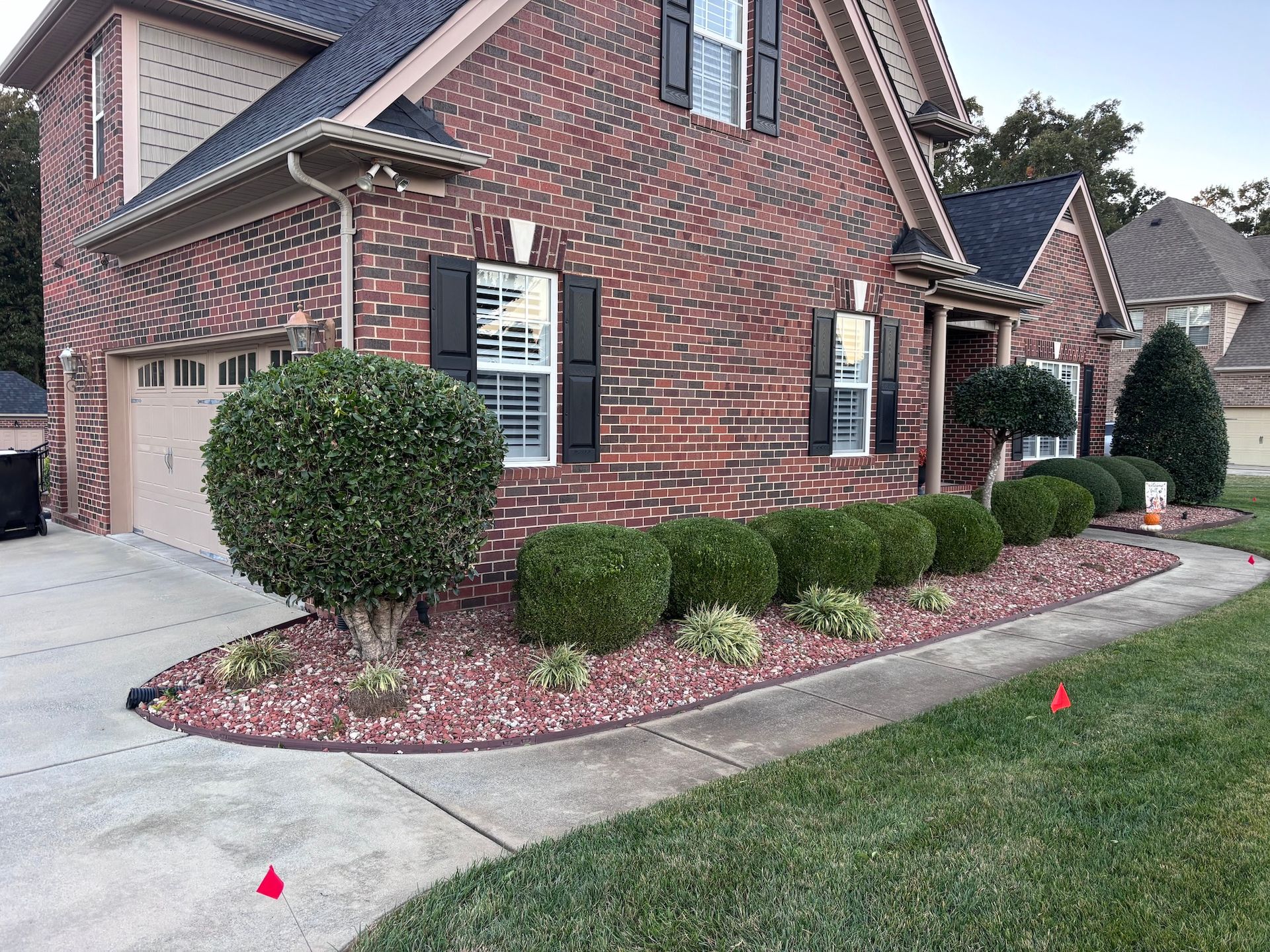 Brick house with black shutters, circular bushes, and a red mulch bed along a sidewalk.