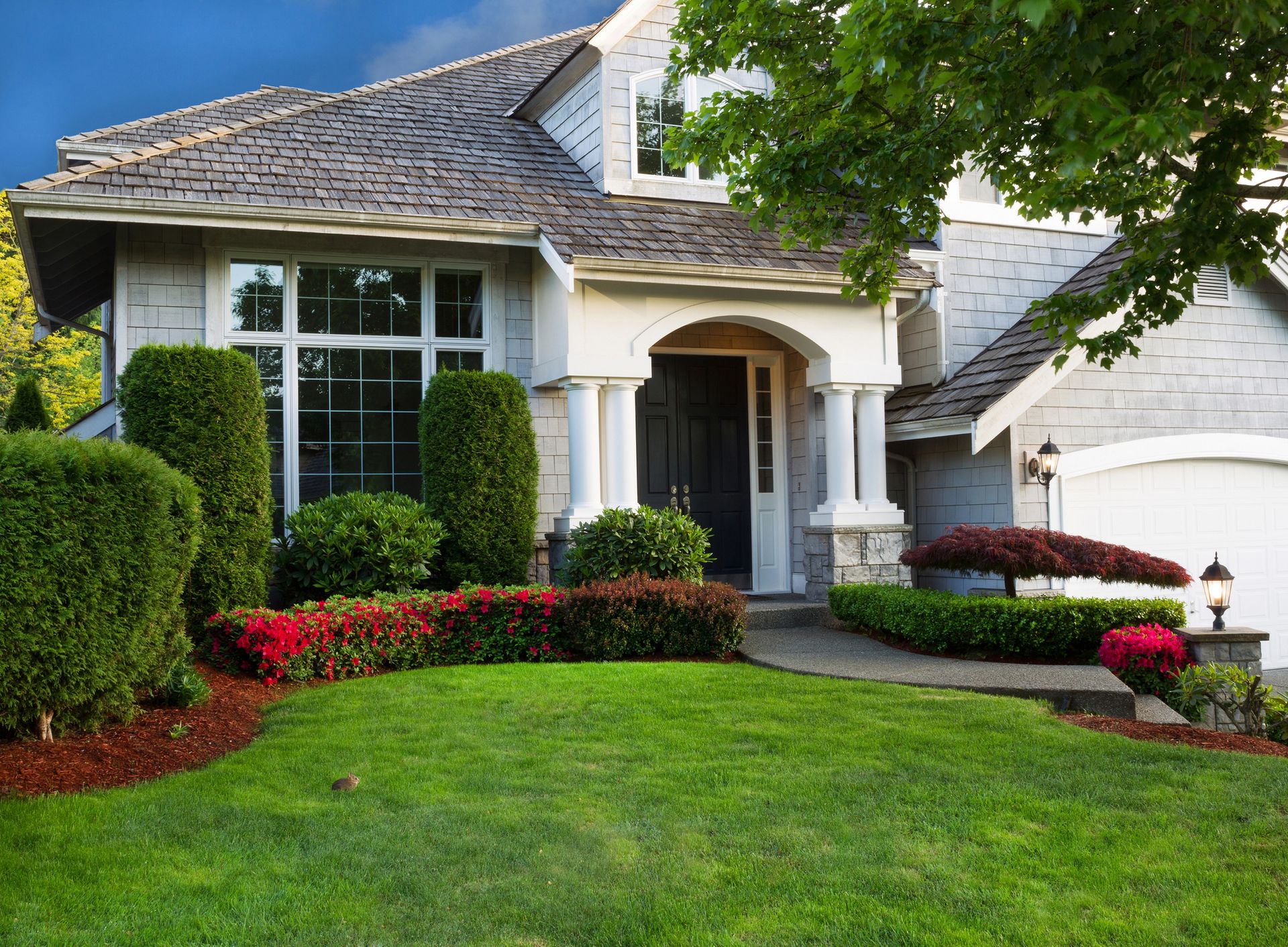 A two-story grey house with white trim and pillars, featuring a neatly landscaped lawn and lush green hedges.