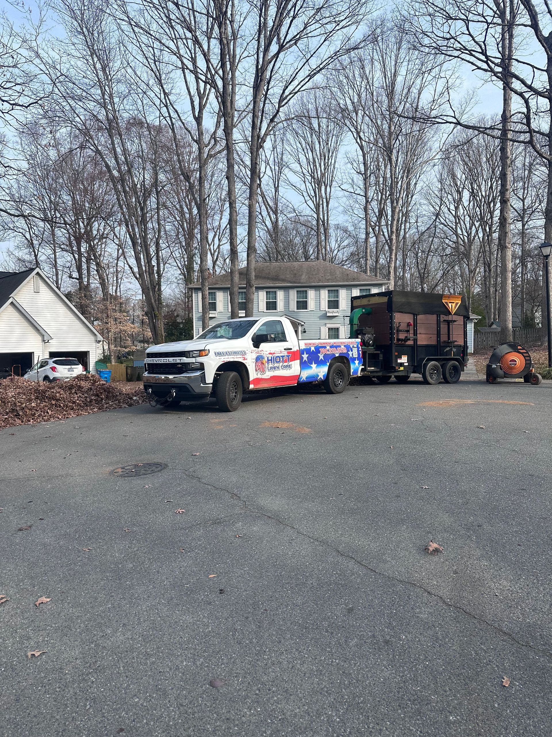 Pile of brown leaves on a street near a house with a lawn, driveway, and leaf blower.