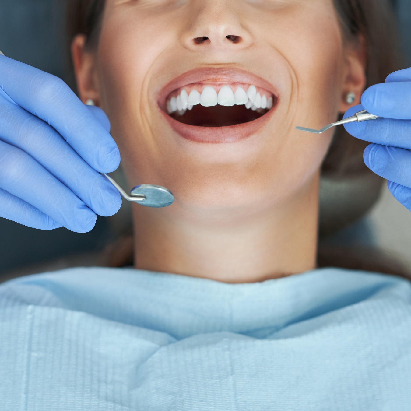 Woman at dentist, mouth open, being examined with instruments by gloved hands.