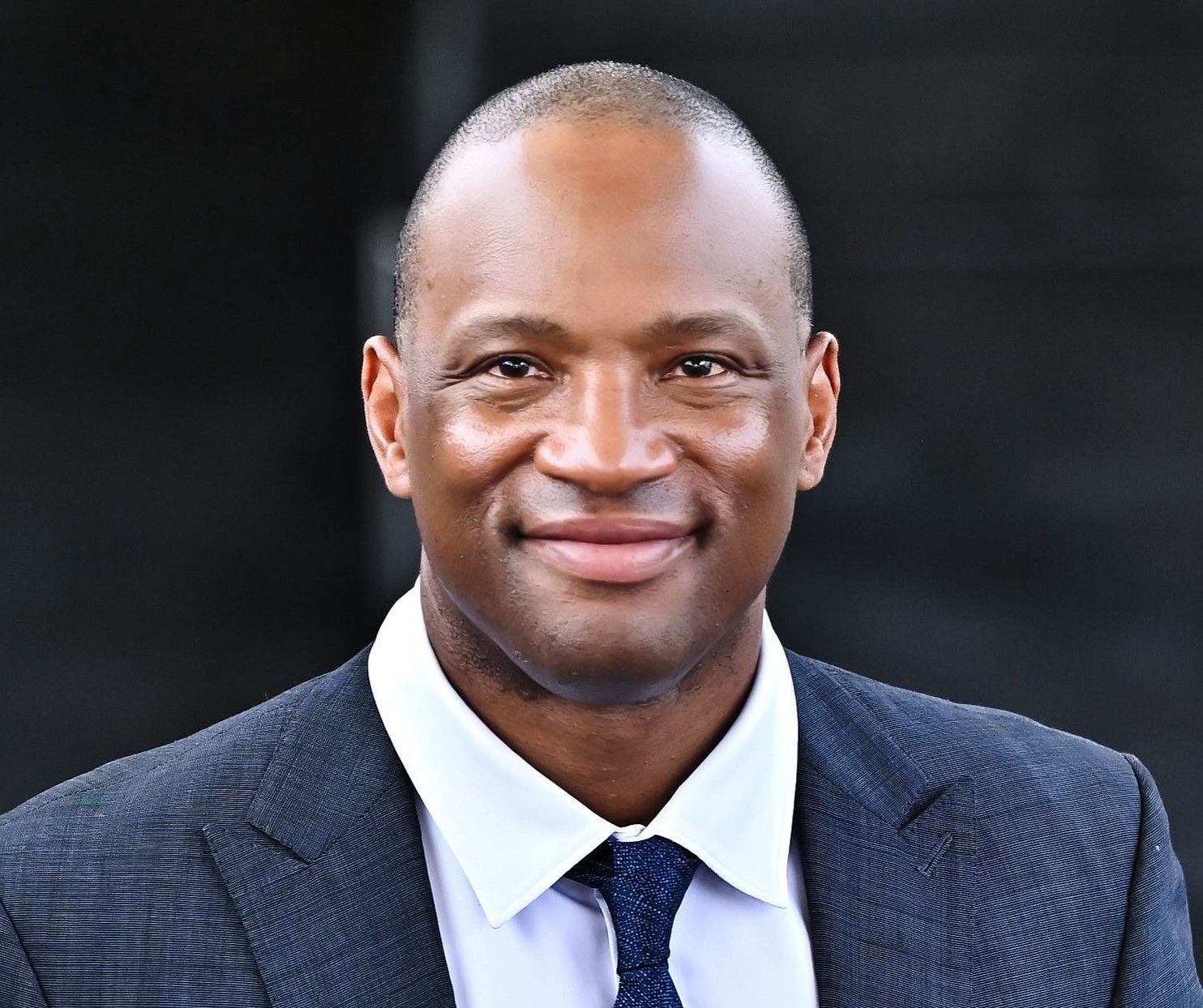 Headshot of a smiling Dane Flanagan wearing a dark grey blazer, white collared shirt, and navy blue tie.