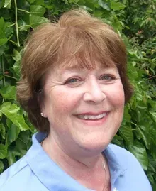 A woman in a blue shirt is smiling in front of a bush.