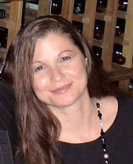 Elaine Scotton with long brown hair smiling at the camera, standing in front of a wine rack.