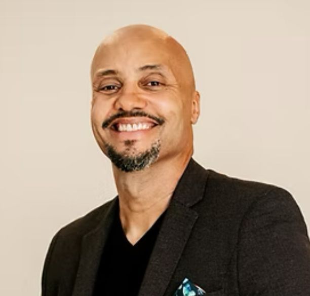 A smiling Richard France  wearing a dark blazer over a black V-neck shirt, against a light background.