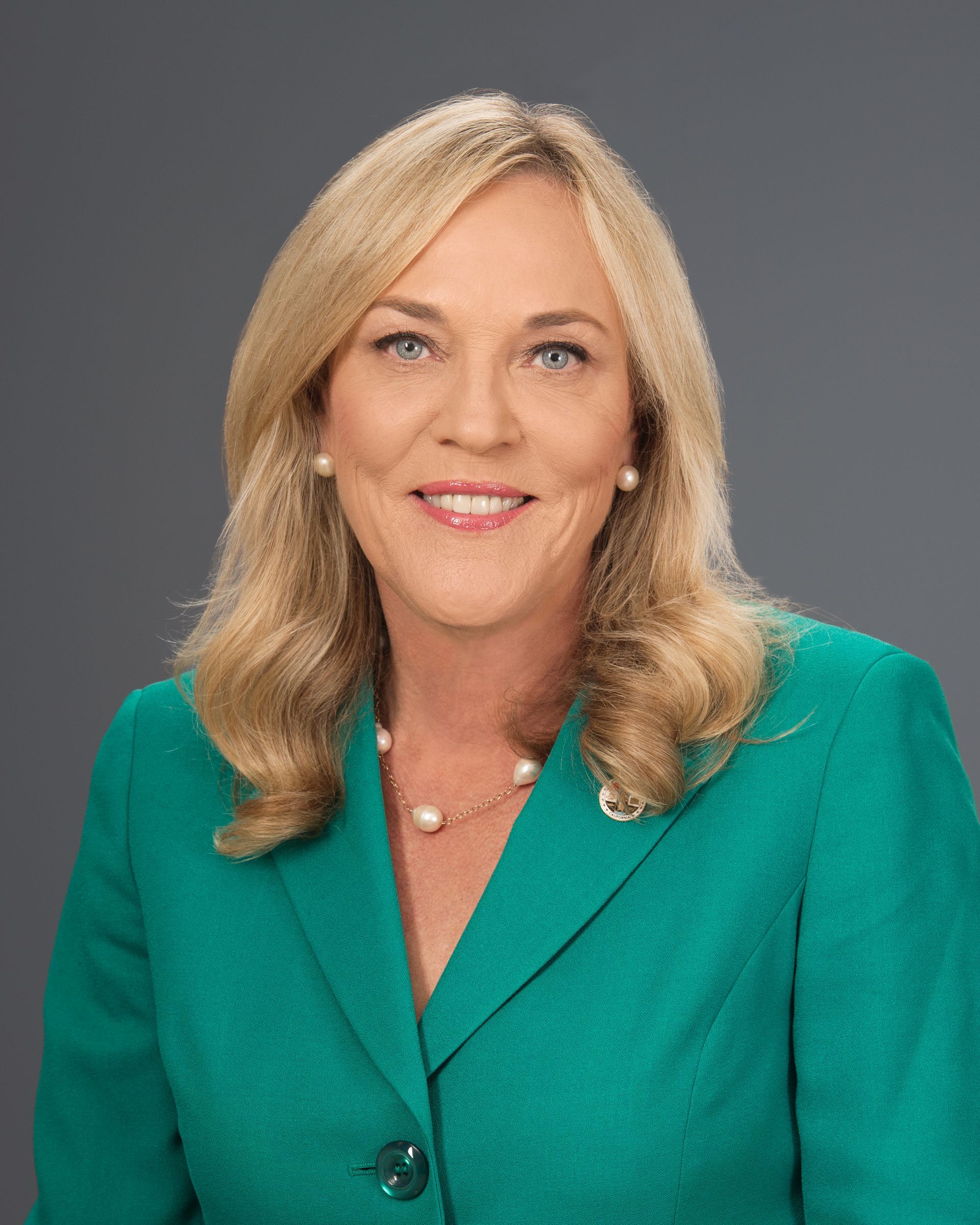A woman in a green jacket and pearl necklace is smiling for the camera.