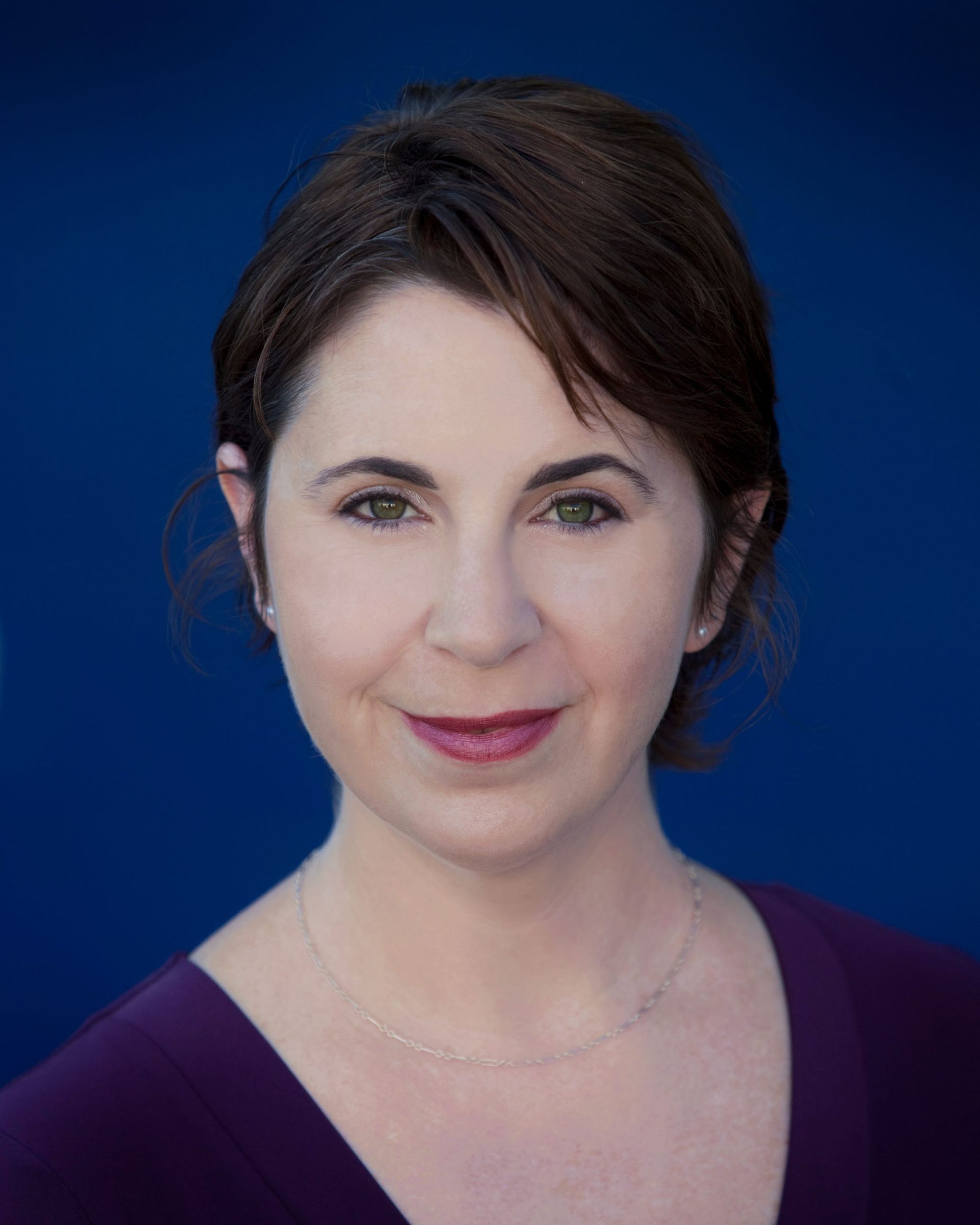 A woman in a purple shirt is smiling in front of a blue background.