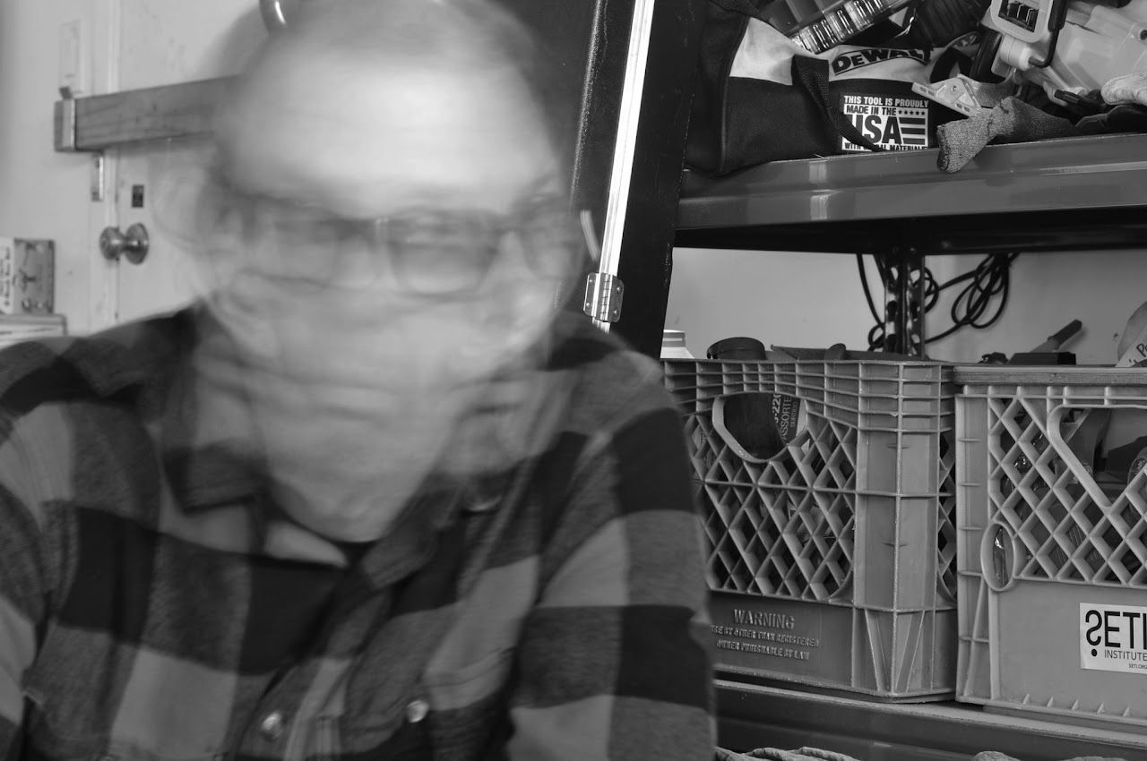 Person in a plaid shirt leaning forward indoors beside stacked crates and shelves.