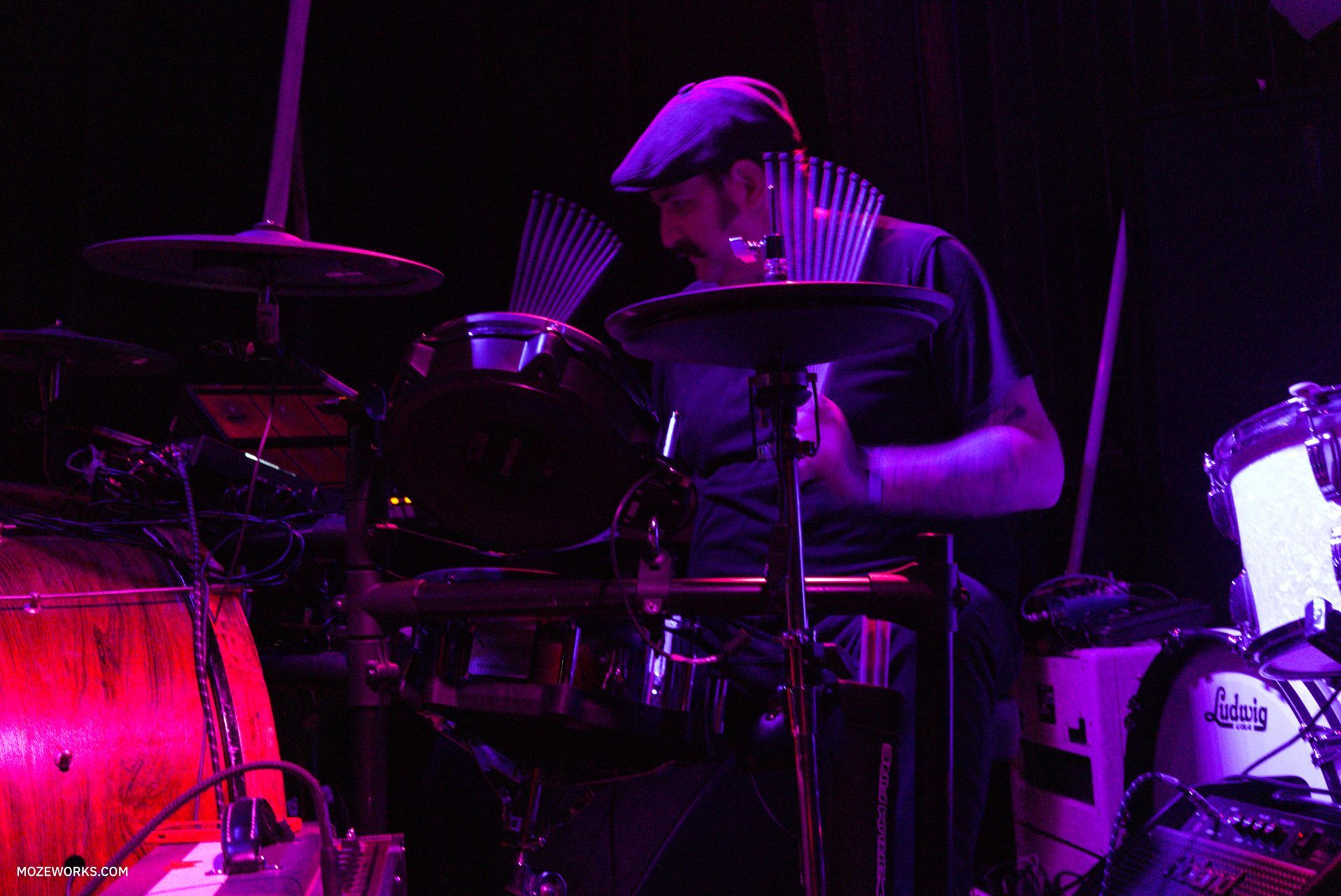 Drummer playing on a dimly lit stage, lit by purple and red lights, surrounded by drum kit cymbals and drums