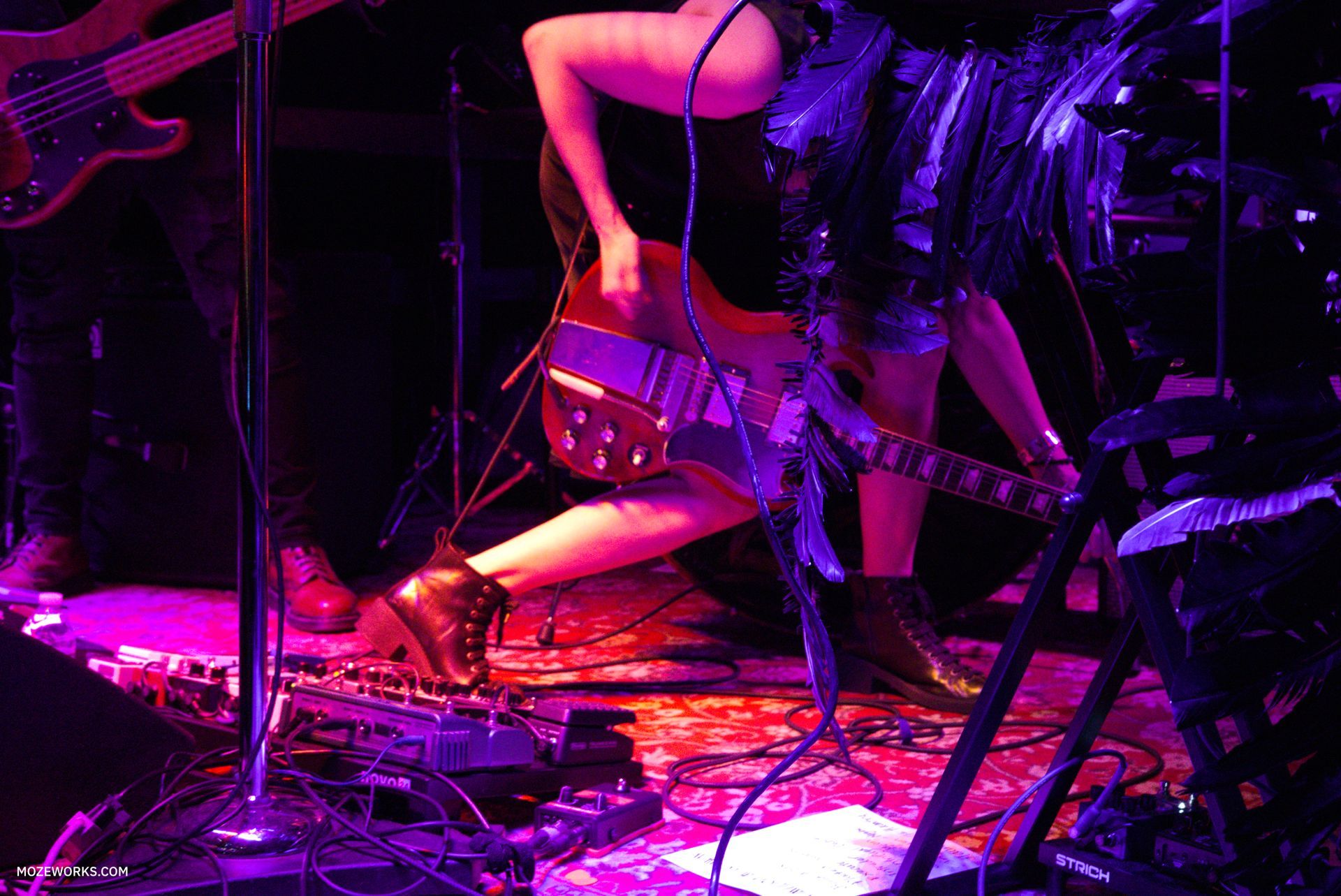 Musician playing guitar on a purple-lit stage with drums and cables visible