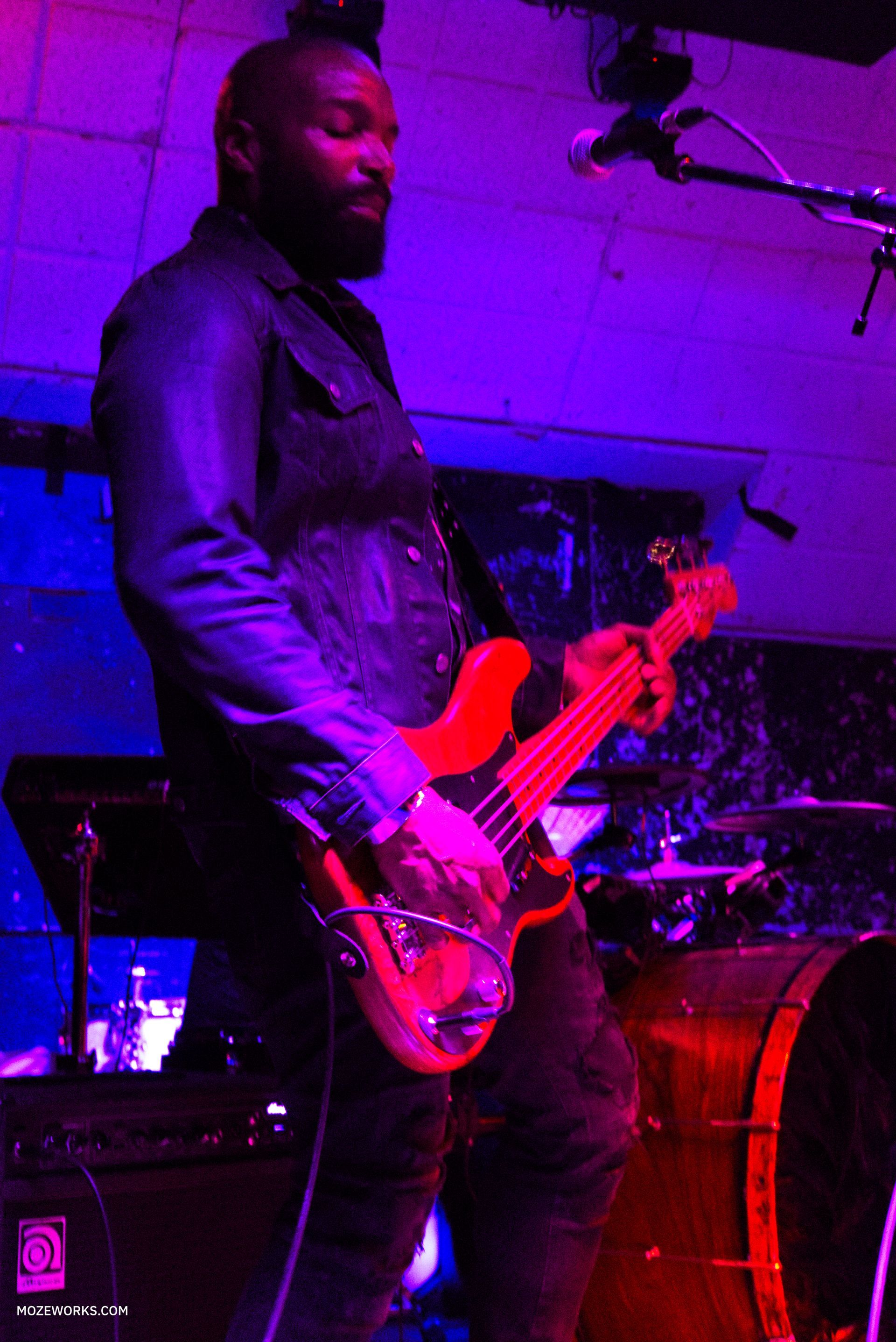 Musician playing a red electric bass on a dimly lit purple stage