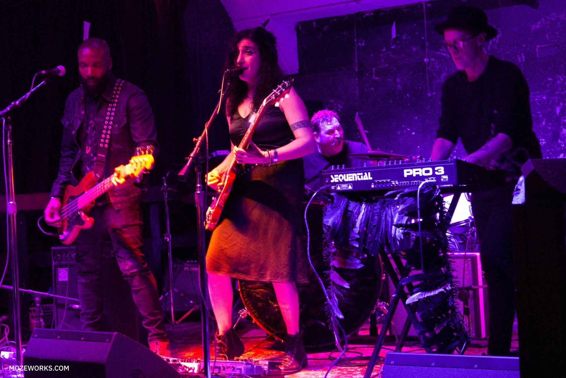 Musicians performing on a purple-lit stage, with guitarists and a keyboard player.
