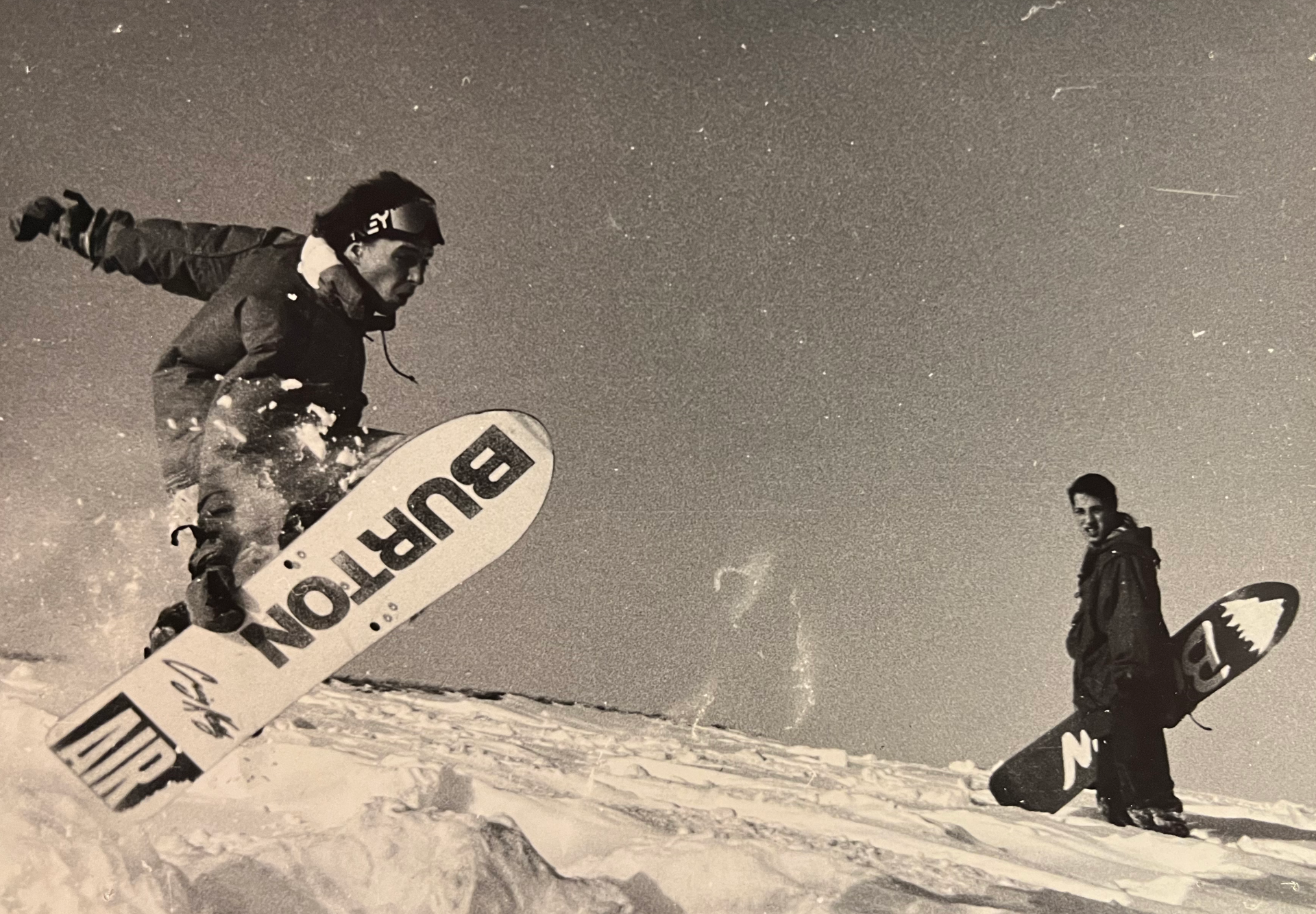 Snowboarder airborne on a snowy slope, with another rider standing nearby.
