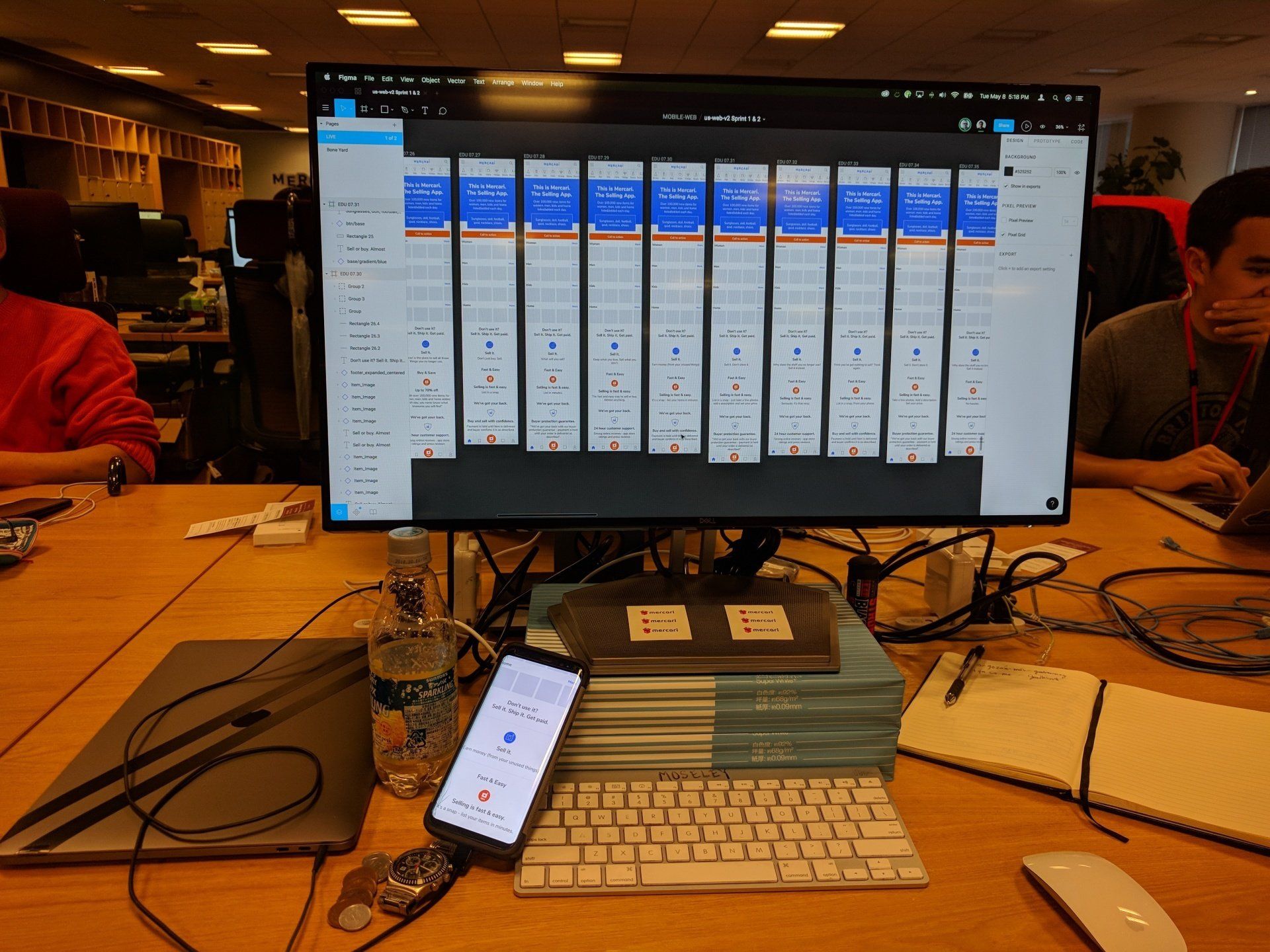 Computer monitor showing a multi-column dashboard on a desk with a phone and keyboard in a busy room