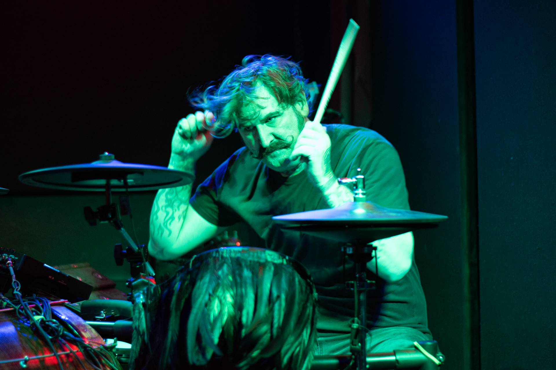 Drummer onstage under green and blue lights, playing intensely behind a drum kit.